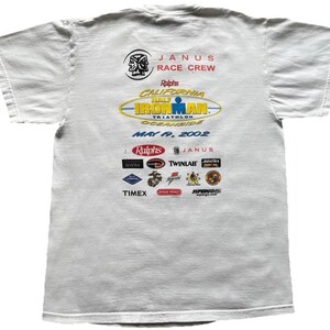 Pu&ograve; includere: Una t-shirt bianca a maniche corte con una stampa grafica sul retro. Il design include il testo JANUS RACE CREW, Ralphs California Half Ironman Triathlon Oceanside May 19, 2002, e vari loghi di sponsor tra cui Timex e Twinlab.