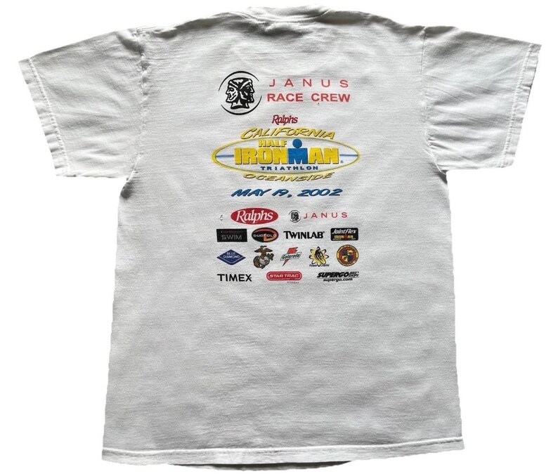 Pu&ograve; includere: Una t-shirt bianca a maniche corte con una stampa grafica sul retro. Il design include il testo JANUS RACE CREW, Ralphs California Half Ironman Triathlon Oceanside May 19, 2002, e vari loghi di sponsor tra cui Timex e Twinlab.