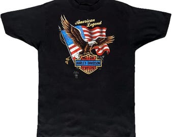 May include: A black short-sleeve t-shirt featuring a graphic of a bald eagle in flight in front of an American flag. Below the eagle is a shield-shaped Harley-Davidson Motor Cycles logo in orange, white, and blue with the text American Legend.