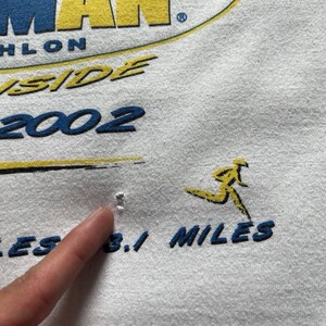 Pu&ograve; includere: Un capo in tessuto bianco con testo serigrafato in blu e giallo che recita MAN, HLON, INSIDE, 2002 e 3.1 MILES, accompagnato da una grafica gialla di una figura che corre. Un piccolo foro circolare &egrave; visibile nel tessuto vicino alla grafica.
