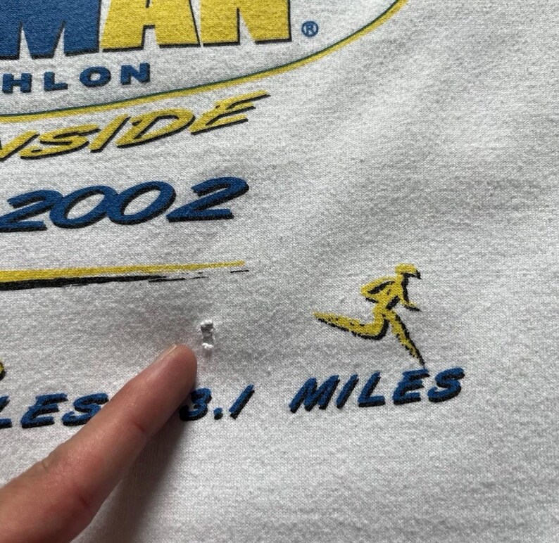 Pu&ograve; includere: Un capo in tessuto bianco con testo serigrafato in blu e giallo che recita MAN, HLON, INSIDE, 2002 e 3.1 MILES, accompagnato da una grafica gialla di una figura che corre. Un piccolo foro circolare &egrave; visibile nel tessuto vicino alla grafica.