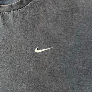 May include: Close-up of a dark gray t-shirt with a white Nike swoosh logo embroidered on the chest. The shirt has a crew neck and a slightly worn appearance, suggesting it's a vintage or pre-owned item.