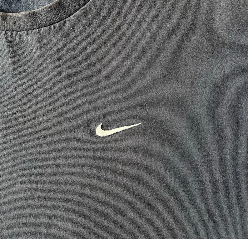 May include: Close-up of a dark gray t-shirt with a white Nike swoosh logo embroidered on the chest. The shirt has a crew neck and a slightly worn appearance, suggesting it's a vintage or pre-owned item.