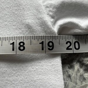 Pu&ograve; includere: Un indumento in tessuto bianco &egrave; posizionato dietro un nastro di misurazione. Il nastro mostra una misura che va da 45,7 a 50,8 cm, con i numeri 18, 19 e 20 chiaramente visibili in nero sullo sfondo bianco dello strumento di misura.