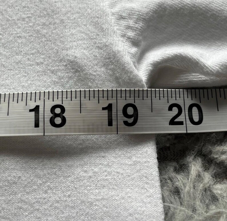 Pu&ograve; includere: Un indumento in tessuto bianco &egrave; posizionato dietro un nastro di misurazione. Il nastro mostra una misura che va da 45,7 a 50,8 cm, con i numeri 18, 19 e 20 chiaramente visibili in nero sullo sfondo bianco dello strumento di misura.