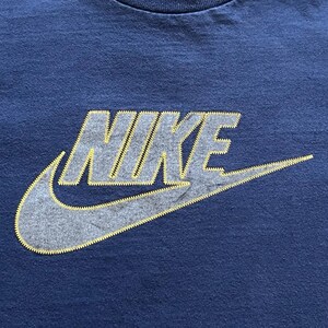 K&ouml;nnte beinhalten: Marineblaues Sport-T-Shirt mit dem Nike-Logo. Das Logo ist grau mit gelben N&auml;hten. Der Nike-Swoosh ist deutlich sichtbar. Das T-Shirt besteht aus einem weichen, bequemen Material.