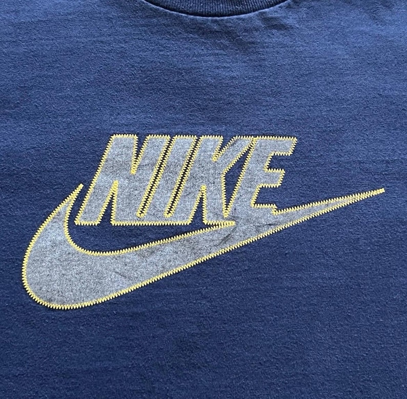 K&ouml;nnte beinhalten: Marineblaues Sport-T-Shirt mit dem Nike-Logo. Das Logo ist grau mit gelben N&auml;hten. Der Nike-Swoosh ist deutlich sichtbar. Das T-Shirt besteht aus einem weichen, bequemen Material.