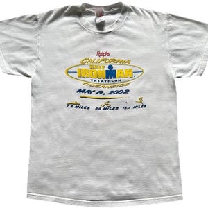 Pu&ograve; includere: Una t-shirt bianca a maniche corte con una grafica per il California Half Ironman Triathlon 2002 a Oceanside. Il design include il testo Ralphs, California Half Ironman, May 19, 2002 e icone per 1,2 miglia, 56 miglia e 13,1 miglia.