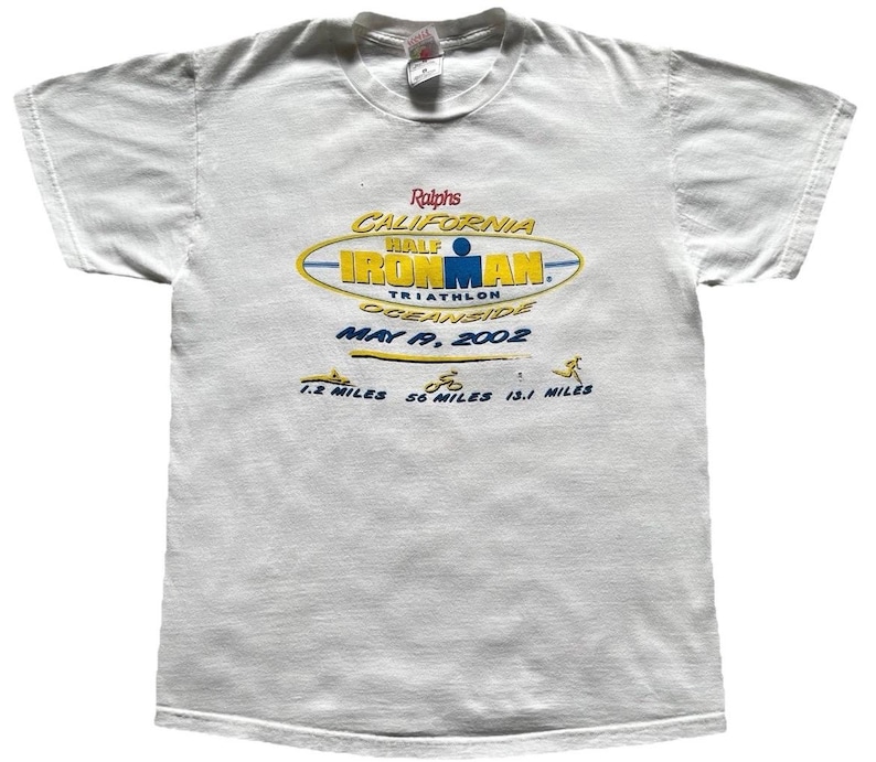 Pu&ograve; includere: Una t-shirt bianca a maniche corte con una grafica per il California Half Ironman Triathlon 2002 a Oceanside. Il design include il testo Ralphs, California Half Ironman, May 19, 2002 e icone per 1,2 miglia, 56 miglia e 13,1 miglia.