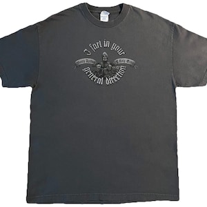 May include: Dark gray t-shirt with a graphic that reads "I fart in your general direction" in a banner. The graphic features a medieval-style illustration. The shirt is a short-sleeved crew neck.