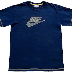 K&ouml;nnte beinhalten: Marineblaues T-Shirt mit goldenem Nike-Logo und Swoosh. Das Shirt hat goldfarbene N&auml;hte an &Auml;rmeln und Saum. Der Kragen ist mit Gold verziert. Kleines Nike-Logo am Kragen.
