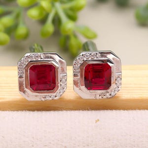 Può includere: Un paio di gemelli quadrati con montatura color argento. Ogni gemello presenta una vivace pietra rossa, taglio quadrato, circondata da un alone di piccole pietre trasparenti. I gemelli sono esposti su una superficie di colore chiaro.