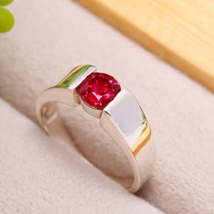 Puede incluir: Un anillo de plata con una llamativa gema roja redonda. El anillo tiene un diseño moderno con un acabado pulido, que resalta la gema. El engaste es de color plata brillante, y la gema es de un rojo intenso.