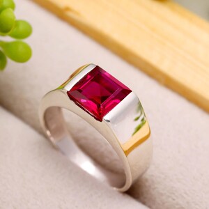 May include: A silver ring with a rectangular, deep red gemstone. The ring has a simple, modern design with a polished finish. The gemstone is set in a bezel setting, and the band is wide and flat. The ring is a statement piece.