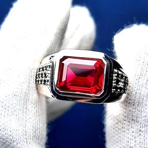 May include: A silver ring with a large, bright red, emerald-cut gemstone. The ring has a textured band with small black accents.