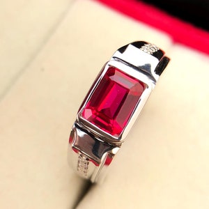 Puede incluir: Un anillo de plata con una piedra preciosa roja engastada en forma cuadrada. El anillo tiene una banda con un diseño decorativo.