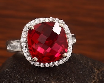 Op de afbeelding: Een zilveren ring met een grote, vierkante rode edelsteen omringd door een halo van kleinere, heldere edelstenen.