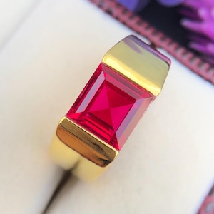 May include: A gold ring with a square, bright red gemstone.