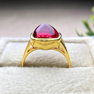 Puede incluir: Un anillo de oro con una gran gema roja oscura en forma de corazón. La gema está engastada en un bisel de oro, y la banda del anillo tiene un diseño delicado y abierto. El anillo se exhibe en una caja de joyería blanca texturizada.