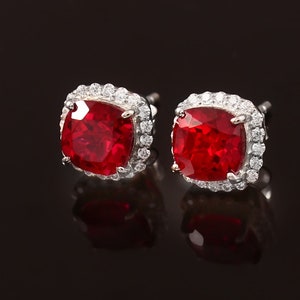 May include: A pair of silver earrings with red square-shaped gemstones surrounded by a halo of smaller clear gemstones.