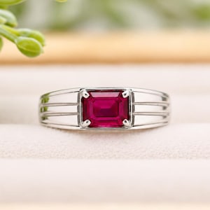 Puede incluir: Un anillo de tono plateado con una piedra preciosa roja vibrante de corte esmeralda rectangular engastada en cuatro garras. La banda presenta tres líneas horizontales ranuradas a cada lado de la piedra central, creando un diseño estructurado.