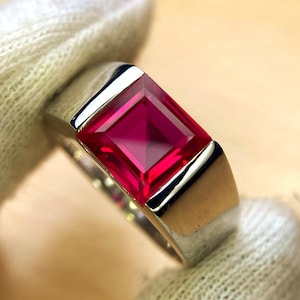 May include: A silver ring with a large, rectangular, faceted ruby. The ruby is a deep red colour, set in a polished silver band. The ring's design is simple but elegant, highlighting the gemstone's brilliance. The ring is a statement piece.