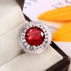 May include: A striking ring featuring a large, round, red gemstone, encircled by a halo of smaller, clear stones. The ring is set in a silver-toned metal and presented in a black box. The design highlights the vibrant red centrepiece.
