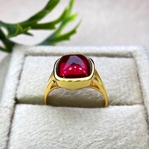 May include: A gold ring featuring a large, square-cut, deep red gemstone. The ring has a classic design with a smooth, polished gold band and a bezel setting that securely holds the vibrant stone. The ring is displayed in a gray jewelry box.