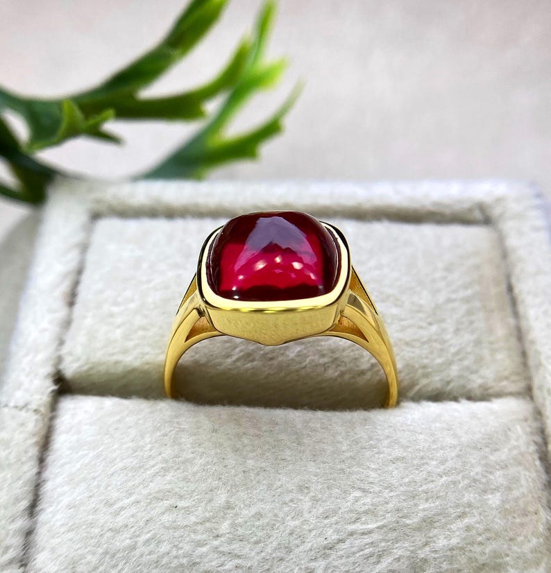 May include: A gold ring featuring a large, square-cut, deep red gemstone. The ring has a classic design with a smooth, polished gold band and a bezel setting that securely holds the vibrant stone. The ring is displayed in a gray jewelry box.