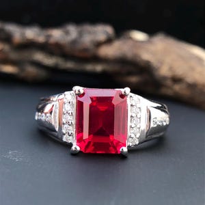 May include: A silver ring featuring a large, rectangular, deep red gemstone as the centerpiece. The ring is accented with smaller, clear gemstones on the band, creating a contrast with the vibrant red. The ring is set against a dark background.