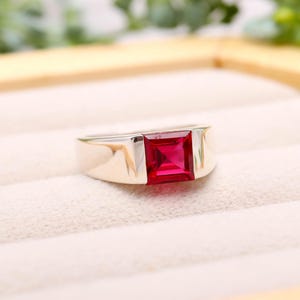 Op de afbeelding: Een zilveren ring met een vierkante, helderrode edelsteen. De ring heeft een eenvoudig, modern ontwerp met een gepolijste afwerking. De edelsteen is gezet in een bezel-zetting en de band is dik en glad.