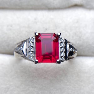 May include: A silver ring featuring a large, rectangular, vibrant red gemstone as the centerpiece. The ring is accented with smaller, clear gemstones on each side of the main stone. The band has a detailed design.
