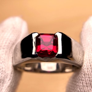 Może przedstawiać: Srebrny pierścionek z dużym, kwadratowym, żywym, czerwonym kamieniem szlachetnym. Pierścionek ma szeroką obrączkę z polerowanym wykończeniem, a kamień jest osadzony w oprawie. Pierścionek jest trzymany przez osobę w białych rękawiczkach.