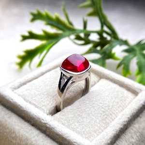 May include: A silver ring with a large, vibrant red gemstone. The gemstone is cushion-cut and set in a bezel setting. The ring's band has a split shank design, adding a modern touch. The ring is displayed on a white, textured surface.