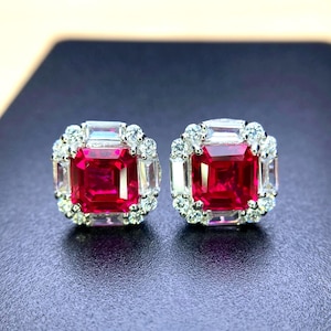 May include: A pair of square-cut ruby earrings with a halo of small, round, and baguette-cut diamonds. The rubies are a deep red color, and the diamonds sparkle against a dark background. The earrings are set in a silver or white metal.
