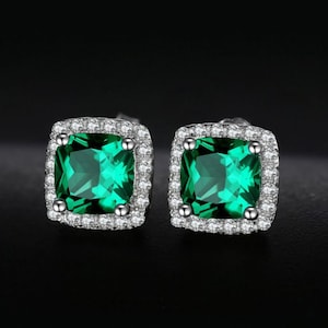 May include: A pair of silver earrings featuring a square emerald green gemstone set within a halo of smaller clear gemstones.