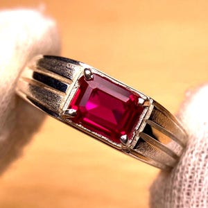 May include: A silver ring with a rectangular, deep red gemstone. The ring has a classic design with a polished band and subtle linear details on the sides, highlighting the gemstone's rich colour and clarity.