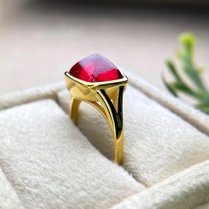 May include: A gold ring featuring a large, triangular, deep red gemstone. The ring has a smooth, polished gold band and a bezel setting that securely holds the vibrant stone. The gemstone's facets catch the light, creating a sparkling effect. The ring is displayed on a soft, white surface.