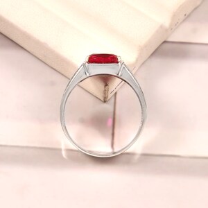 May include: A silver ring with a large, red, square-shaped gemstone. The ring has a simple band with a small, decorative detail on the side.