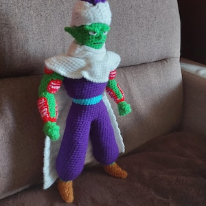 Puede incluir: Una figura de crochet verde y morado con un turbante blanco, brazos a rayas rojas y blancas y una capa blanca. La figura está de pie en un sofá marrón.