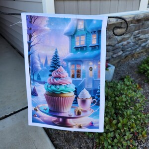 Op de afbeelding: Een decoratieve tuinvaandel met een winterse scène met een cupcake en een huis. De cupcake heeft roze en blauwe glazuur. Een klein kopje slagroom en een geschenkdoos staan ook op de vlag. Het huis is blauw met verlichte ramen.