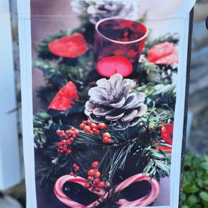Puede incluir: Una bandera de jardín decorativa con un arreglo festivo. El diseño incluye una vela roja, piñas, bayas rojas y bastones de caramelo. El fondo tiene ramas de hoja perenne y acentos rojos, creando un tema navideño.