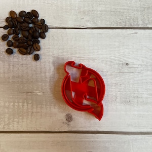 Op de afbeelding: Een rode plastic koekjesvorm in de vorm van een gestileerde letter "M" in een cirkel, met een kleine hoop koffiebonen linksboven. De vorm ligt op een licht houten oppervlak.