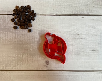 May include: A red plastic cookie cutter in the shape of a stylized letter "M" inside a circle, with a small pile of coffee beans to the upper left. The cutter is on a light wood-grain surface.