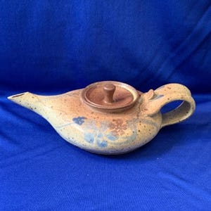 May include: A ceramic teapot with a brown lid and handle, and a spout. The teapot has a speckled beige and brown finish with blue floral accents. The teapot is on a blue background.