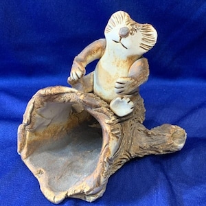 May include: A ceramic koala bear figurine sitting on a hollowed-out log. The koala is light brown and cream-colored, with detailed facial features. The log is textured with a natural, earthy brown color. The figurine is on a blue background.