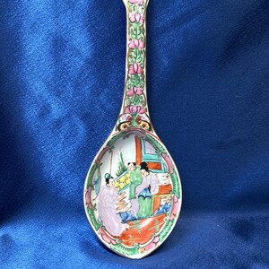 Puede incluir: Una cuchara decorativa de porcelana con un mango largo y una cabeza en forma de cuenco. El mango está adornado con diseños florales rosas y hojas verdes. El cuenco presenta una escena colorida de figuras en una habitación, con detalles en rojo, verde y blanco.