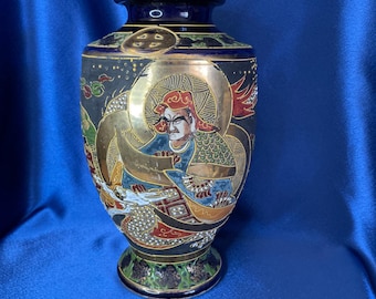 May include: A decorative, hand-painted vase with a dark blue rim and base. The vase features a detailed illustration of a figure with a golden halo and accents. The background is black with gold stars and the vase is approximately 6 inches tall.