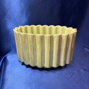May include: A pale green ceramic bowl with a scalloped edge. The bowl has vertical ridges and a glossy finish. The bowl is a light green colour and sits on a blue surface.