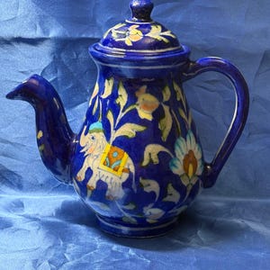 May include: A cobalt blue ceramic teapot featuring a hand-painted design of a white elephant with a yellow and orange saddle, surrounded by white and light blue floral motifs. The teapot includes a matching lid with a small knob handle.
